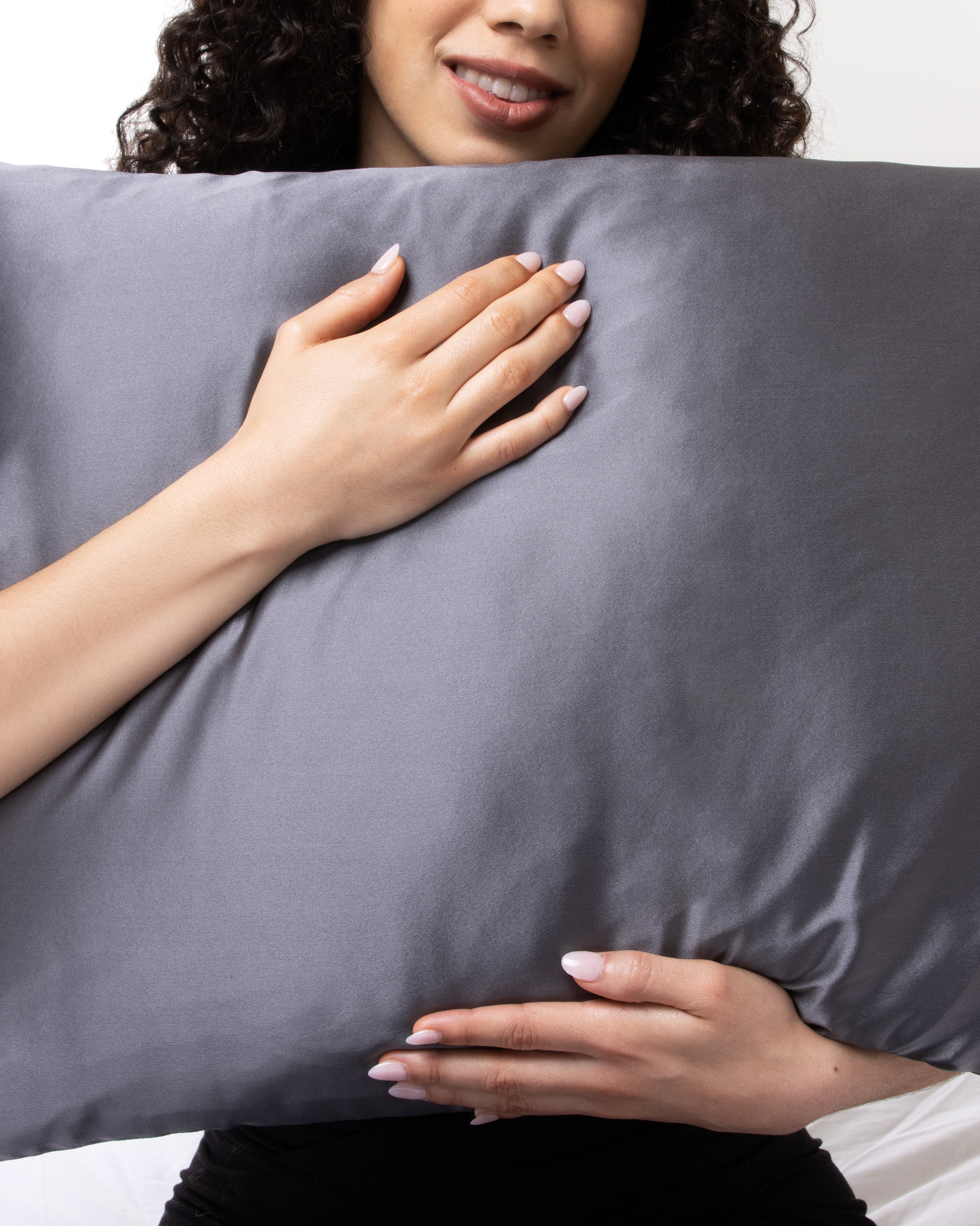 A woman holding a dark grey silk pillowcase against her chest, showing the smooth sheen of the fabric.