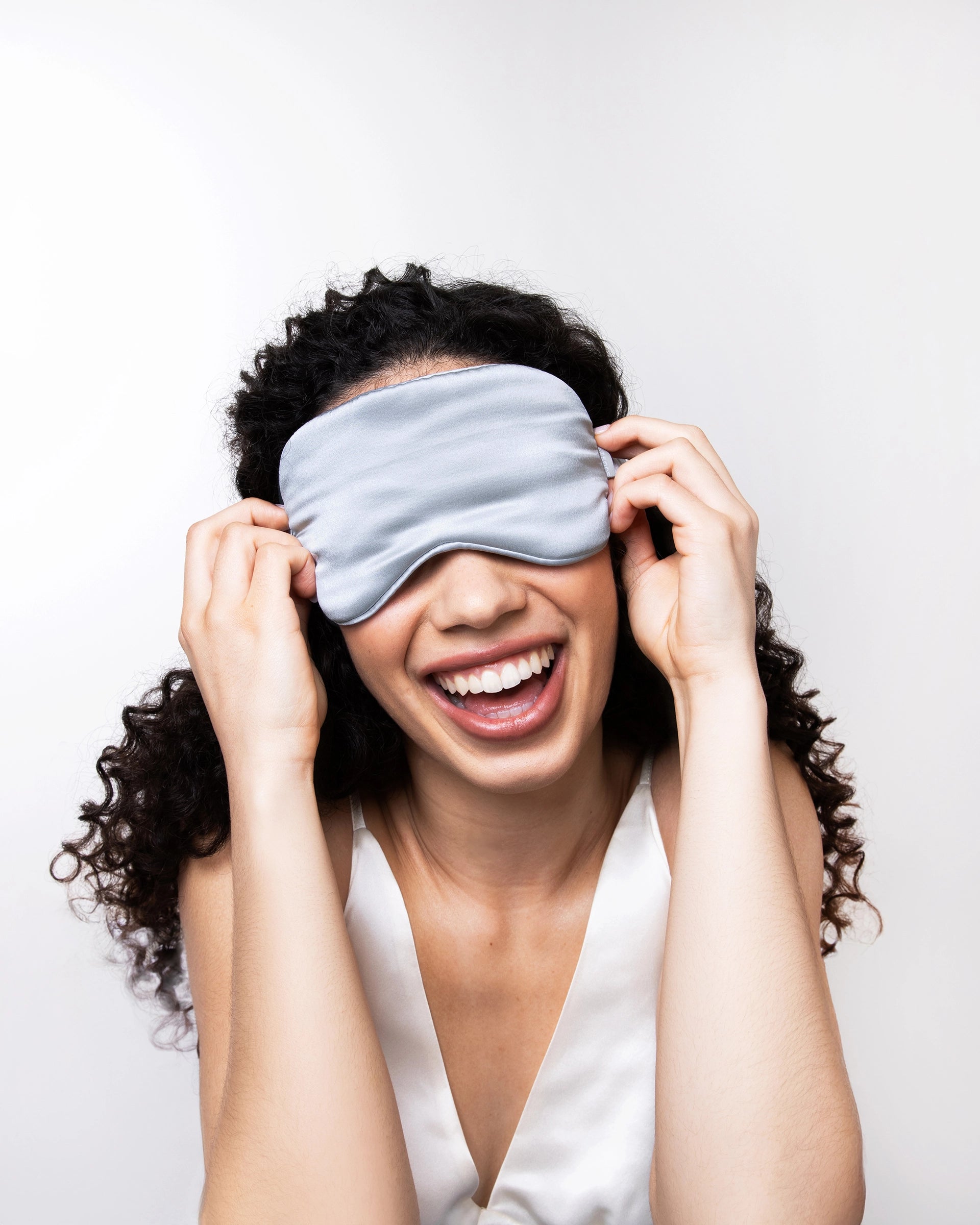 A person with curly hair laughs while pulling a smooth, light-grey silk sleep mask over their eyes, creating a playful, relaxed mood